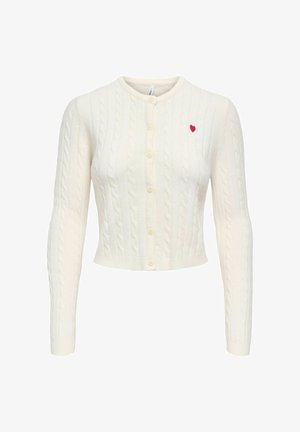 Bela skrajšana jopica z vzorcem pletenja v obliki kablov, z dolgimi rokavi, okroglim izrezom, sprednjimi gumbi in majhnim rdečim srcem izvezenim na levi strani prsi.