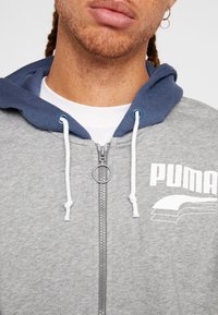 Haut de sweat à capuche gris zippé avec une capuche à cordon bleu, des cordons blancs et un logo à l'avant. Il présente un tissu texturé et une fermeture éclair métallique.