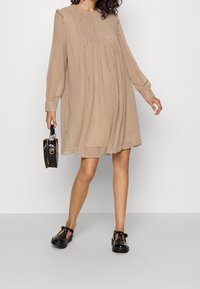 Beige Kleid mit gepunktetem Muster, langen Ärmeln und gerafftem Oberteil; kombiniert mit einer schwarzen Handtasche und glänzenden schwarzen T-Riemen-Schuhen.