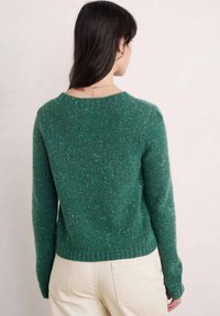 Zelený svetr s tečkovaným vzorem, dlouhými rukávy a kulatým výstřihem. Ribkované lemy a manžety. Nosič má na sobě světlé kalhoty.