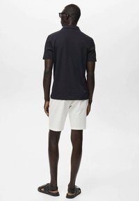 Svart polotröja, ljusa shorts och sandaler. Kort ärm, enkel design och avslappnad passform. Bakifrån mot en vit bakgrund.