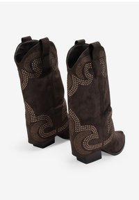 Botas de vaquero de ante marrón con un diseño de tachuelas patrón, con un tacón bajo y lengüetas en la parte superior para facilitar su uso.