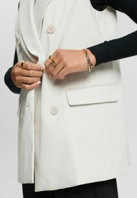 Weißer ärmelloser Blazer mit Doppelreiher-Design, zwei Fronttaschen und glatter Textur; die Hände des Modells geschmückt mit goldenen Ringen und Armbändern.
