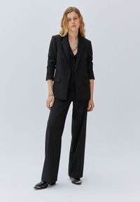 Traje de rayas negras que incluye un blazer ajustado y pantalones de pierna ancha. El blazer tiene mangas enrolladas y se combina con un chaleco de botones.