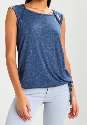 Kobieta ubrana w bezrękawnikowy niebieski top w kropki z guzikami na ramieniu i wiązaniami po bokach, w połączeniu z jasnoniebieskimi spodniami.