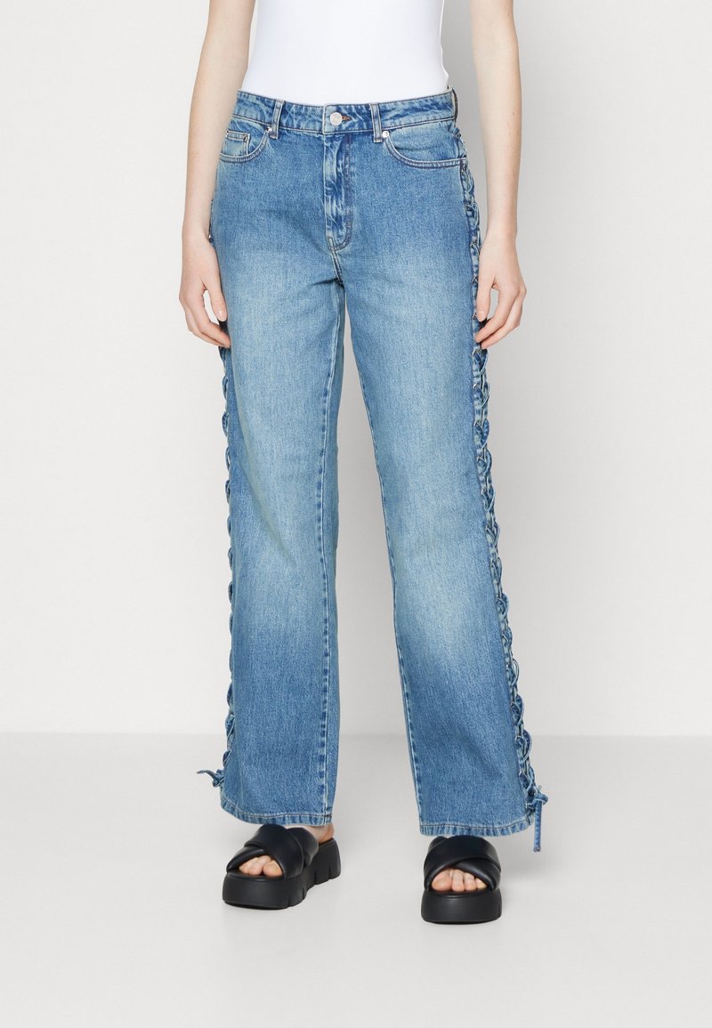 Jeans de mezclilla de pierna ancha en azul claro con detalles de encaje a lo largo de los costados. Cuenta con bolsillos frontales y cierre de botón, combinados con sandalias negras gruesas.