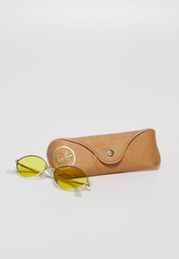 Gafas de sol Ray-Ban con lentes amarillos, montura metálica y puntas de patillas transparentes. Incluye un estuche beige texturizado con logo dorado y cierre a presión.