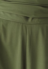 Tela verde oliva con textura suave, con una pretina doblada y costuras verticales que le dan una apariencia estructurada.