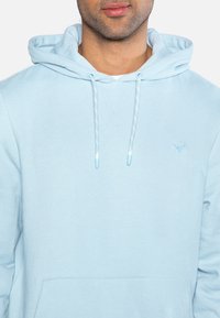 Světle modrá mikina s kapucí vyrobená z měkké tkaniny, s klokaní kapsou, šňůrkami a malým vyšitým motivem na hrudi.