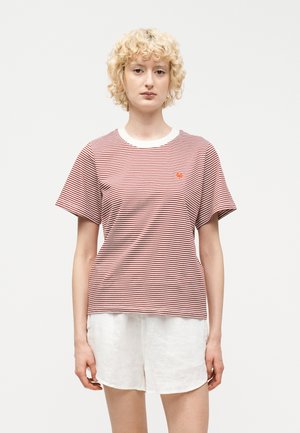 Junge Person mit lockigem blondem Haar, trägt ein rot-weiß gestreiftes T-Shirt und weiße Shorts, steht vor einem einfarbigen hellen Hintergrund.