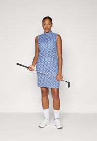 Modré golfové šaty bez rukávov so vzorovaným dizajnom, s vysokým golierom a priliehavým strihom. Model drží golfovú palicu a má na sebe biele ponožky a topánky.