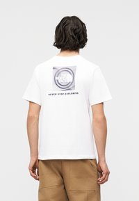 Άτομο με σκούρα μαλλιά μέχρι τους ώμους που φοράει λευκό μπλουζάκι με κυκλικό γραφικό και το κείμενο «ΠΟΤΕ ΜΗ ΣΤΑΜΑΤΑΣ ΝΑ ΕΞΕΡΕΥΝΑΣ» στην πλάτη.