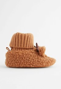 Bruna stickade tofflor med en texturerad, mjuk kropp, en ribbad manschett och en dekorativ detalj. Sulan är platt och slät.