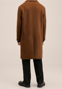 Manteau en laine marron à coupe droite, col cranté et épaules tombantes, associé à un pantalon noir et des chaussures.