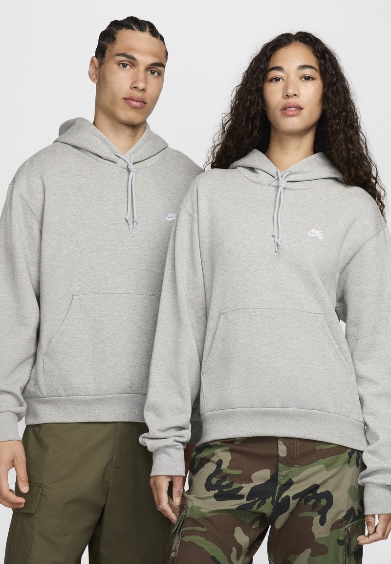 Sivi bombažni puloverji s kapuco z žepom spredaj, zategovalnim vrvico in majhnim belim logotipom Nike. Modeli nosijo cargo hlače z vzorcem kamuflaže.