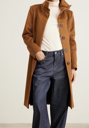 Henkilö pukeutuneena kamelinväriseen pitkään takkiin, valkoisen pooloneuleen ja tummansinisten leveälahkeisten farkkujen kanssa, toinen käsi taskussa.