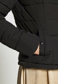 Giacca a vento nera con design trapuntato, colletto arrotondato e tasca laterale con chiusura a scatto. La texture appare liscia e leggermente lucida.