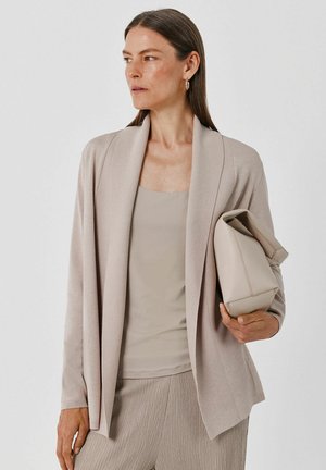 Woman with long brown hair wearing beige cardigan, tank top, and pleated pants holding matching beige handbag, looking to side.