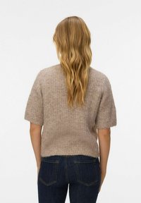 Femme aux cheveux blonds ondulés portant un pull en tricot beige à manches courtes et un jean bleu foncé, vue de dos sur fond blanc.