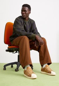 Man sitter på en orange kontorsstol iförd en mönstrad mörkgrön tröja, bruna byxor, vita strumpor och tan Adidas sneakers på ett ljusgrönt golv.