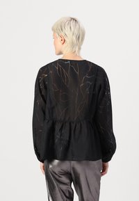 Svart chiffonblus med peplumdesign, långa ärmar och ett lövmönster i subtila färger. Mjuk textur och genomskinligt utseende.