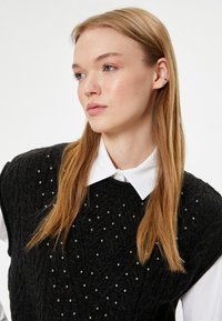 Schwarze, gestrickte ärmellose Sweater mit strukturiertem Muster, verziert mit kleinen Metallic-Perlen, getragen über einem weißen Hemd mit Kragen.