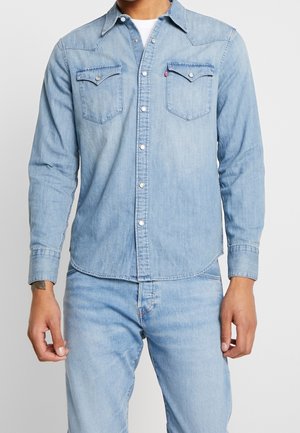Homme portant une chemise en jean bleu clair avec boutons-pression, poches poitrine, et un jean en jean bleu clair assorti, debout devant un fond uni.