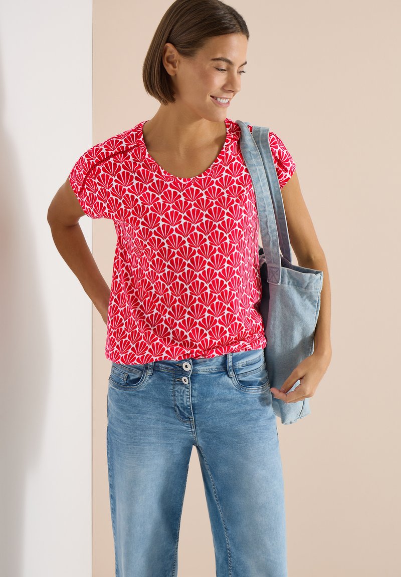 Jeune femme portant une chemise rouge à motifs floraux et un jean bleu, avec un sac fourre-tout bleu clair sur l'épaule, souriant en regardant vers le bas.