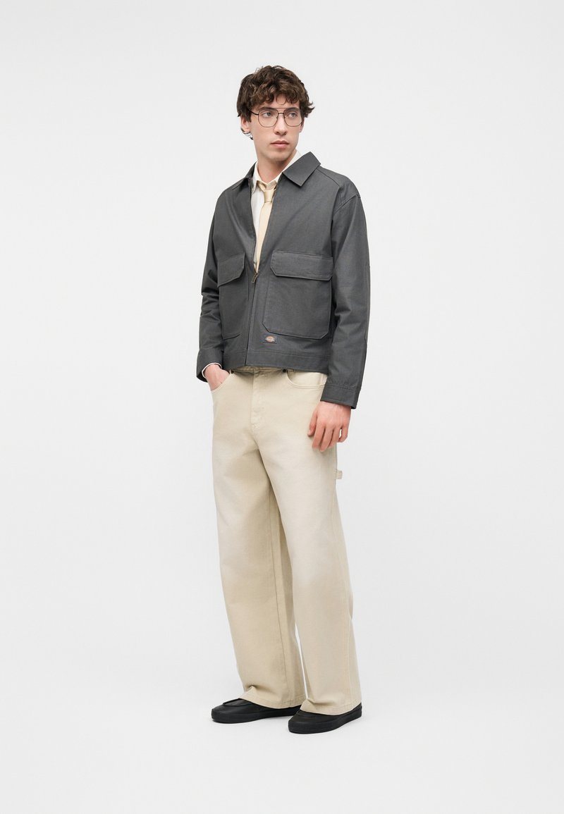 Jeune homme portant une veste grise zippée, un pantalon beige à jambes larges, des chaussures noires et des lunettes, debout les mains dans les poches sur fond blanc.