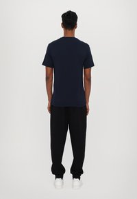 Námořnická modrá tričko s krátkým rukávem, bavlněný materiál, kulatý výstřih, uvolněný střih, nošeno s černými kalhotami a bílými teniskami, pohled zezadu.