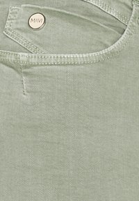 Denim verde con una textura sutil, con una costura cosida y un botón de metal dorado grabado con "MAVI". Detalle del bolsillo visible.