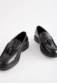 Mocassins en cuir noir avec pampilles, présentant une finition lisse, un détail tressé sur la tige et un talon bas et robuste.