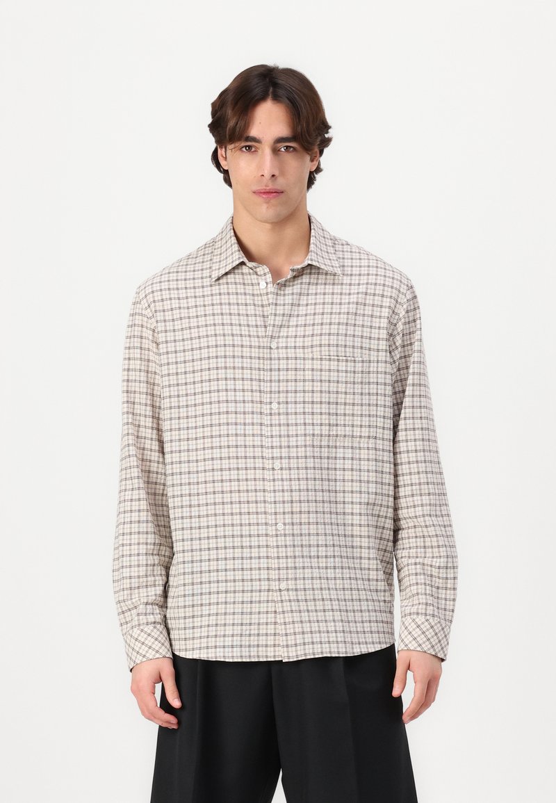 Camisa de manga larga, con botones, en un tejido marrón claro con un patrón cuadrado en marrón oscuro y beige. Cuenta con un bolsillo en el pecho y cuello puntiagudo.