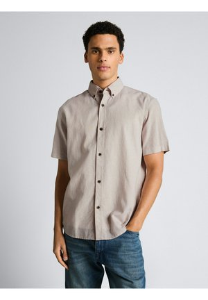 Junger Mann steht mit einer Hand in der Tasche, trägt ein hellbeiges kurzärmeliges Hemd und blaue Jeans vor einfarbigem Hintergrund.