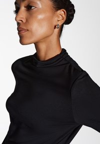 Schwarzes Oberteil mit hohem, gefälteltem Kragen, das aus glattem, figurbetontem Stoff gefertigt ist und eine elegante Silhouette hat. Silberne Ohrringe setzen einen dezenten Akzent.