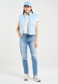 Šviesiai mėlynas pūkų liemenė, be rankovių, su aukštu apykakle, uždėta ant baltos marškinėlių. Suderinta su nusidėvėjusiais šviesiai mėlynais džinsais ir pilkais sportbačiais.