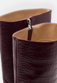Bottes hautes en cuir bordeaux embossé crocodile avec un intérieur lisse, comprenant une fermeture éclair argentée à l'ouverture supérieure pour un enfilage facile.
