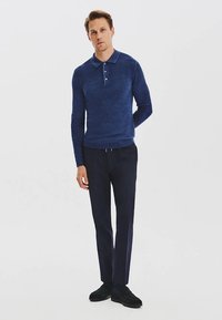 Granatowa koszulka polo z teksturowanej tkaniny, z zapięciem na trzy guziki oraz klasycznym kołnierzykiem, zestawiona z ciemnymi spodniami i czarnym obuwiem.