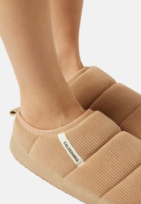 Beige slip-on tøfler med et tekstureret, ribbet design, med en blød stofoverdel og et kontrasterende hvidt mærke på siden.