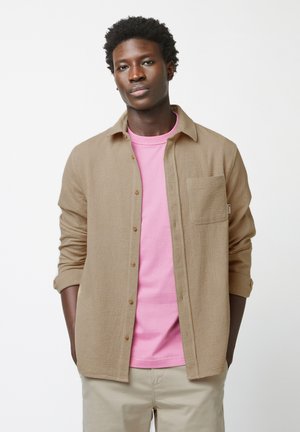 Camicia button-up color beige in tessuto strutturato sopra una t-shirt rosa, con taschino sul lato sinistro del petto, maniche arrotolate e un taglio casual.