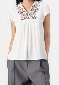 Blouse blanche avec broderie florale de couleurs vives, manches courtes volantées et coupe décontractée avec un design plissé.