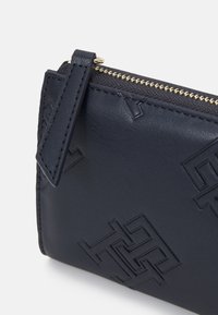 Compatto portamonete in pelle blu navy caratterizzato da motivi geometrici in rilievo, completato da una cerniera dorata e un'anello laterale per un facile trasporto.
