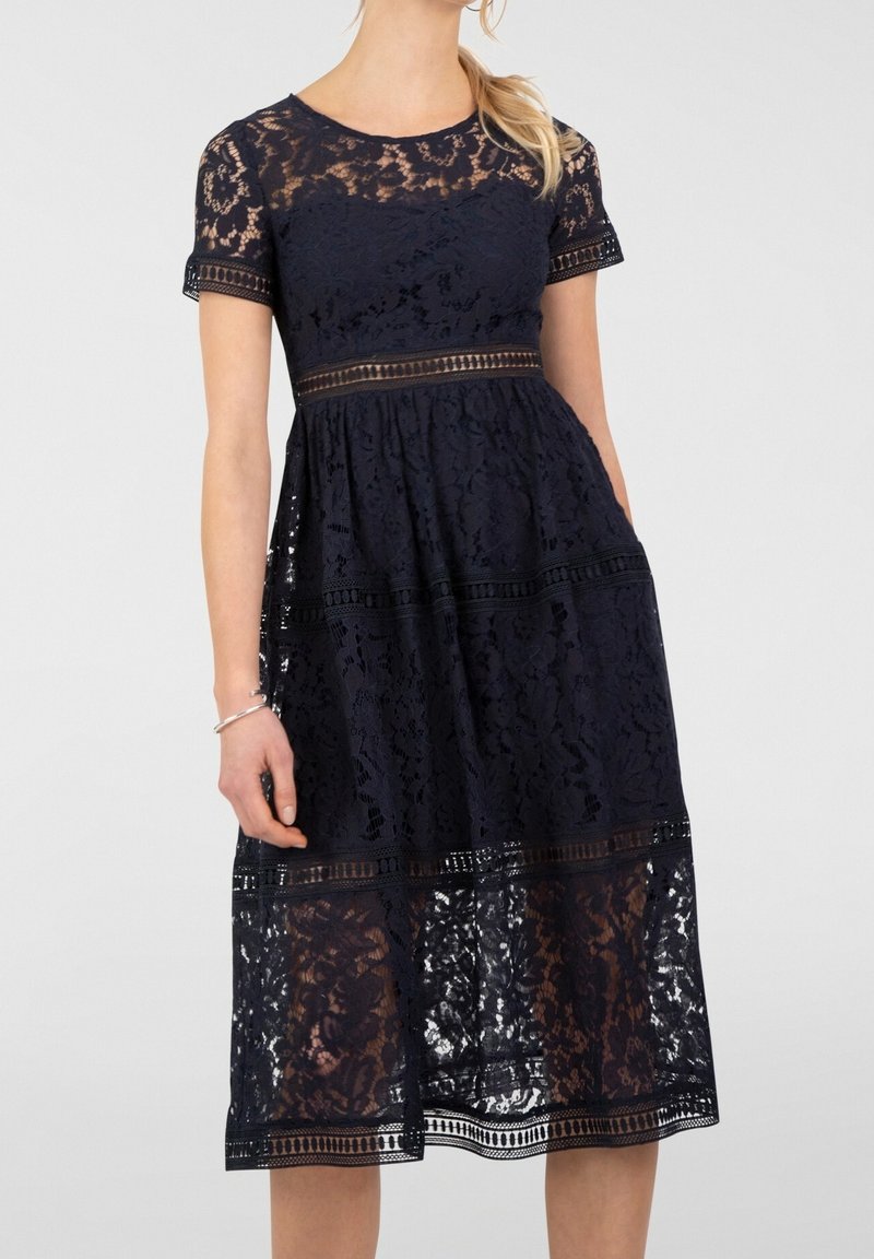 Femme portant une robe midi en dentelle bleu marine avec des manches courtes, des sections transparentes au niveau du décolleté, de la taille et de l'ourlet, debout devant un fond uni.