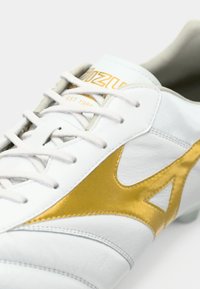 Nogometni čevelj iz belega usnja z zlatimi poudarki, teksturirano površino in tradicionalnim zapiranjem z vezalkami, z zlatim logotipom na jeziku.