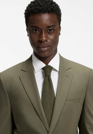 Mann trägt eine olivgrüne Anzugjacke, weißes Hemd und eine strukturierte olivgrüne Krawatte vor einem schlichten weißen Hintergrund.