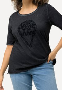 Černé tričko s krátkým rukávem s texturovanou výšivkou ve tvaru srdce, s kulatým výstřihem a volným střihem. V kombinaci se světlými modrými džíny.