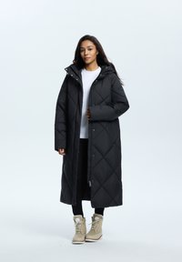 Schwarzer, steppierter langer Mantel mit Kapuze, ausgestattet mit einem Reißverschluss vorne, Seitentaschen und einer glatten Textur. Trägt einen weißen Top und schwarze Hosen.