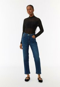 Schwarzes langärmliges Mesh-Oberteil mit hohem Kragen, kombiniert mit blauen geraden Jeans aus Denim mit vorderen Taschen und Knopfverschluss. Schwarze Ballerinas.