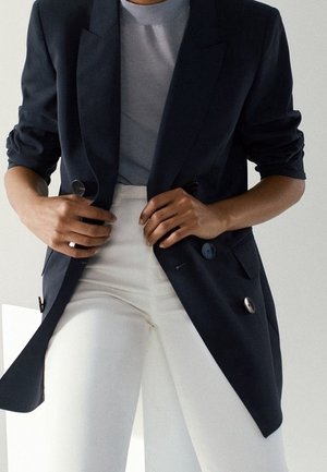 Person wearing a navy blazer over a gray shirt and white high-waisted pants, adjusting the blazer with both hands.