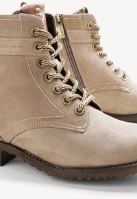 Bottines beige en daim à lacets avec surpiqûres contrastées, œillets ronds en métal, fermeture éclair latérale et semelle en caoutchouc marron texturée.